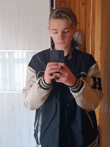 a young man taking a picture of himself in a mirror wearing a varsity jacket with the letter h on the sleeves