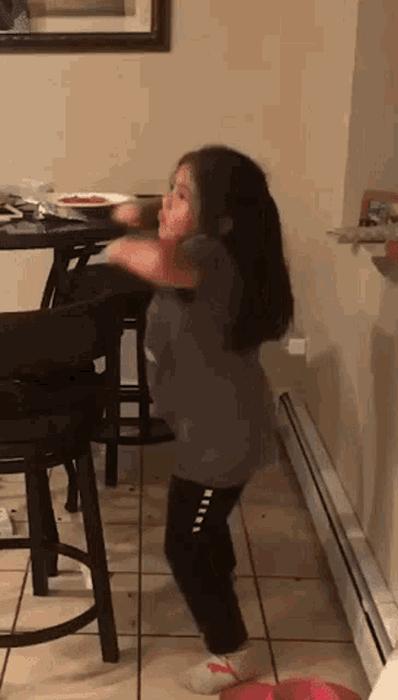 a little girl is dancing in a kitchen in front of a table and stools .