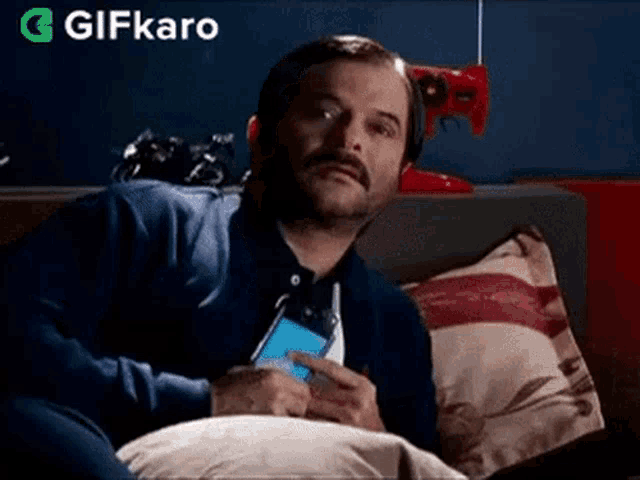 a man with a mustache is sitting on a couch holding a cell phone in his hand .