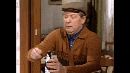 a man in a brown sweater and hat is sitting at a table holding a bottle of beer .