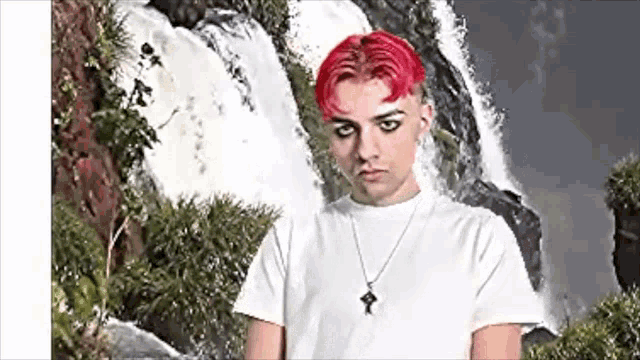 a young man with red hair is standing in front of a waterfall wearing a white shirt and a necklace .