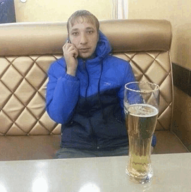 a man in a blue jacket is talking on a cell phone next to a full glass of beer