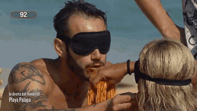 a man wearing blindfolds is eating noodles from a woman on the beach