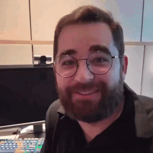 a man with glasses and a beard smiles in front of a computer monitor