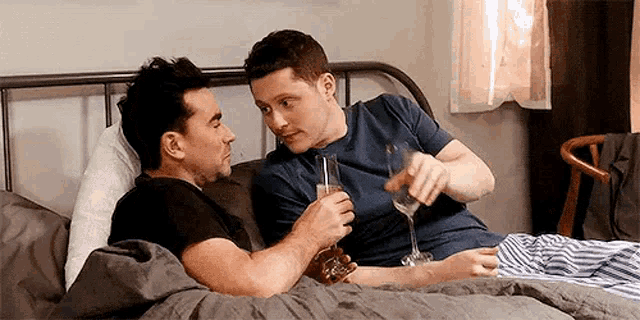 two men are laying on a bed holding glasses of champagne .