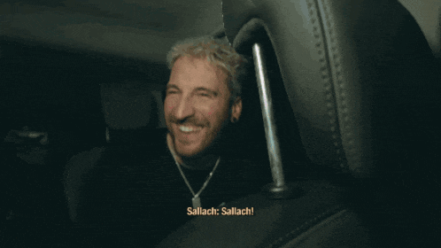 a man with a beard is smiling in a dark room with the words sallach below him