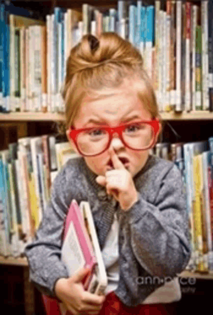 a little girl wearing red glasses is holding a book in front of a bookshelf