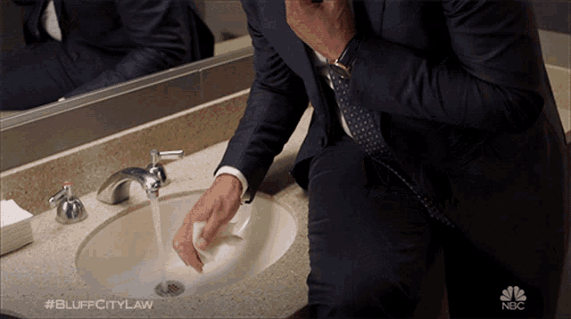 a man in a suit is washing his hands in a bathroom sink with nbc written on the bottom