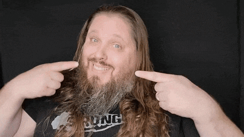 a man with long hair and a beard is pointing to his face .