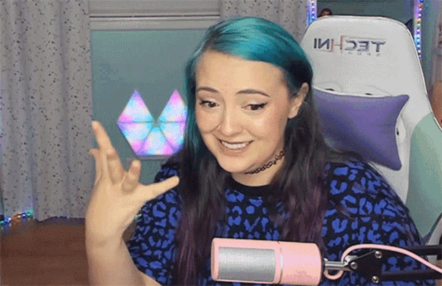 a woman with blue hair stands in front of a pink microphone with the word techi on the back of the chair