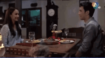a man and a woman sit at a table with plates of food and a clock in the background