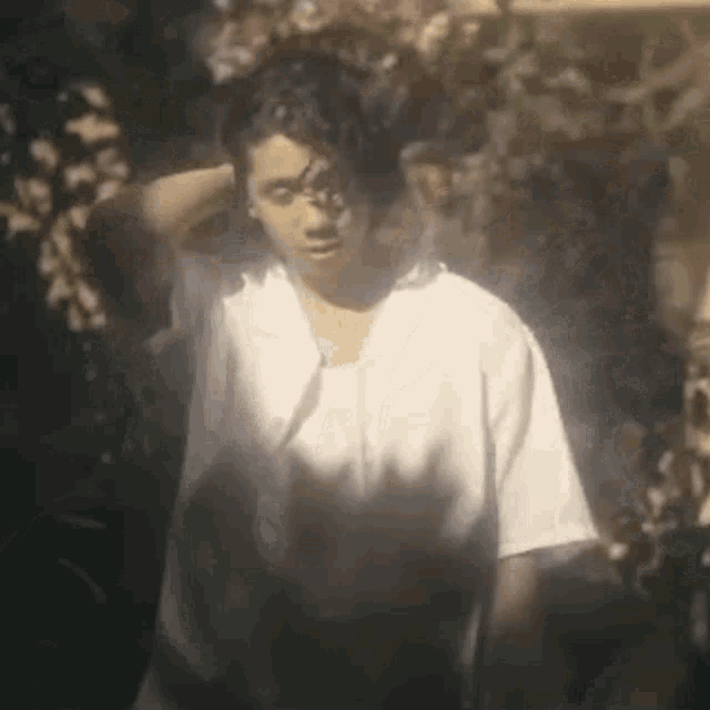 a man in a white shirt is standing in the shade of a tree with his hand on his head .