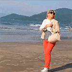 a woman in red pants is standing on a beach holding a white purse .