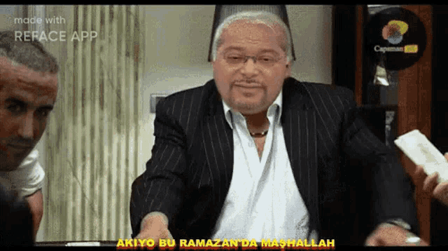 a man in a suit and white shirt is standing in front of a sign that says akiyo bu ramazan da mashallah