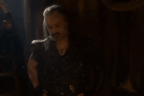 a man with long hair and a beard is standing in a dark room holding a knife .