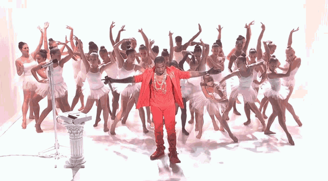 a man in a red suit stands in front of a microphone surrounded by ballerinas