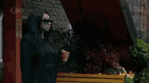 a woman wearing sunglasses and a black hoodie stands in front of a brick building