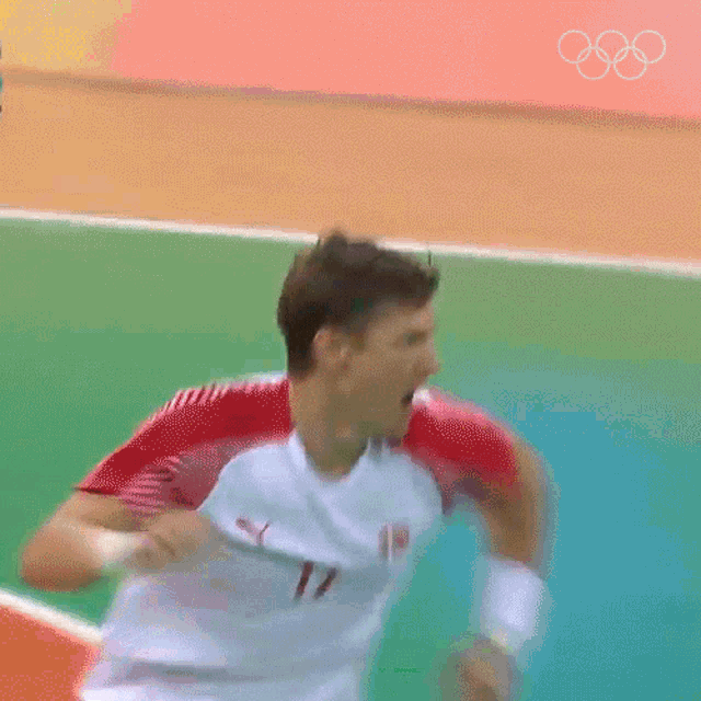 a man in a red and white shirt is running on a green field with the olympic rings in the background .