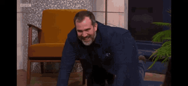 a man in a blue shirt is crawling on the floor in front of a yellow chair with bbc written on the bottom