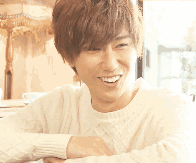 a young man wearing a white sweater smiles with his arms crossed