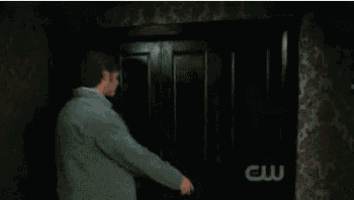 two men are standing in a dark room with a cw logo on the wall behind them