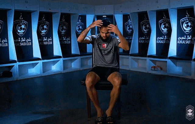 a man sits in a chair in a locker room that says alhilal saudi club on the wall