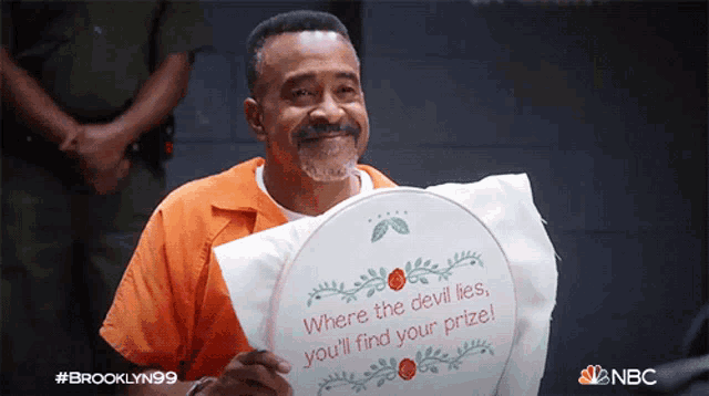 a man in an orange shirt holds a sign that says where the devil lies you 'll find your prize