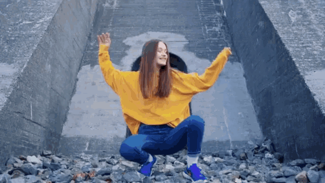 a woman wearing a yellow sweater and blue jeans is squatting on a pile of rocks with her arms outstretched .