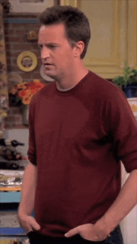 a man in a red shirt is standing in a kitchen with his hands in his pockets .