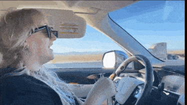 a woman wearing sunglasses is driving a car with a warning label on the rear view mirror