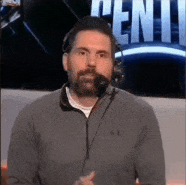 a man with a beard wearing headphones and a under armour shirt is talking into a microphone .