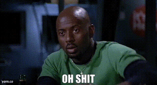 a bald man in a green shirt is sitting at a table with the words oh shit written on his face .