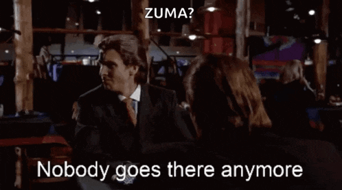 a man in a suit and tie is talking to a woman in a dark room and says zuma