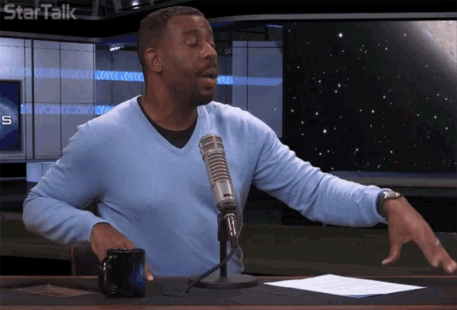a man in a blue sweater stands in front of a microphone with the word startalk behind him