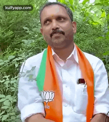 a man wearing a white shirt and an orange and green scarf is standing in the woods with his arms crossed .