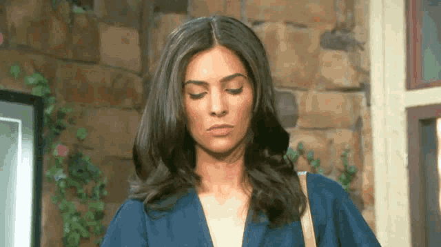a woman in a blue dress is standing in front of a stone wall and looking down .