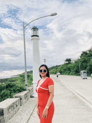 a woman in a red dress and sunglasses stands in front of a lighthouse