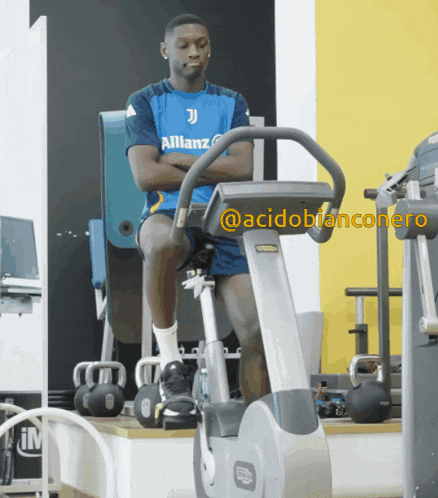a man in a blue allianz shirt sits on an exercise bike in a gym