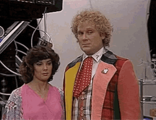 a man and a woman are standing next to each other in a room . the man is wearing a pink suit and tie .