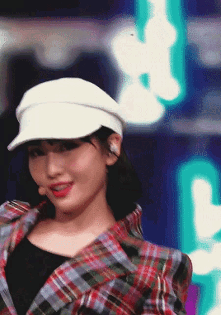 a woman wearing a plaid jacket and a white hat looks at the camera