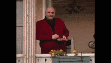 an elderly man in a red sweater is standing on a balcony holding a plate of food .