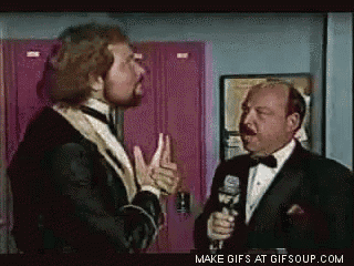 a man in a tuxedo is talking to another man with a microphone .
