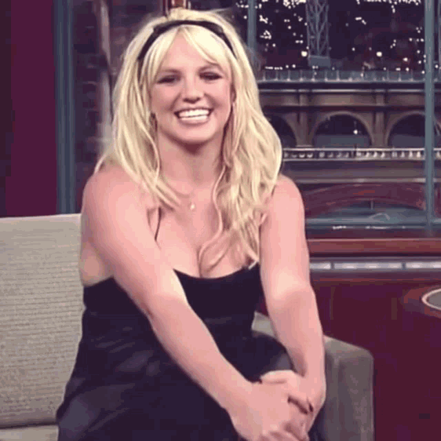 a blonde woman in a black dress is sitting on a couch and smiling