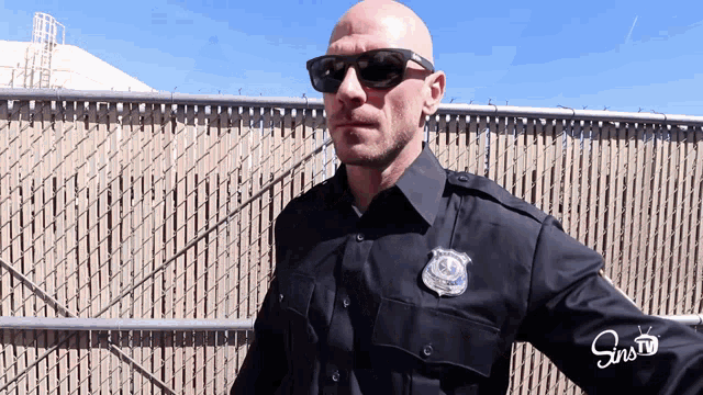 a bald police officer wearing sunglasses stands in front of a fence with sins tv written on his sleeve