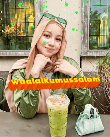 a woman wearing a hijab sits at a table with a drink and a sign that says waalaikumusalam