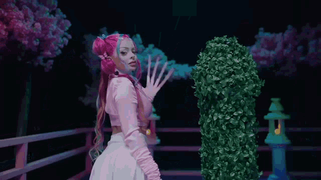 a girl with pink hair and long nails is standing in front of a bush .