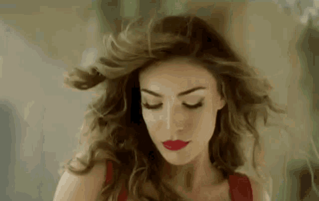 a woman with long hair and red lipstick is standing with her eyes closed and her hair blowing in the wind .