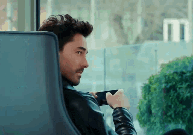 a man in a leather jacket is sitting in front of a window holding a cell phone
