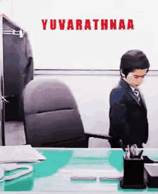 a man in a suit and tie is standing in front of a desk with the words yuvarathaa on the wall behind him .