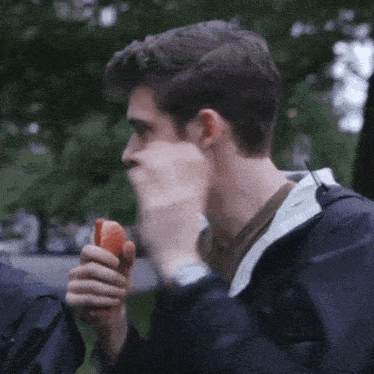 a man in a black jacket is eating a hot dog in a park